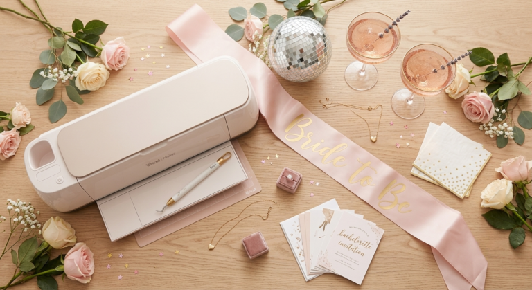 A top-down flat lay photograph on a light wood surface, showcasing a rose and champagne gold Cricut Maker 3 and crafting tools surrounded by bachelorette party decor. A pink satin sash is draped across the scene with the text "BRIDE TO BE" in readable gold foil lettering. Nearby are rose gold champagne coupe glasses, pink and cream roses, eucalyptus sprigs, a disco ball, and bachelorette invitation cards in velvet boxes. The composition highlights a cohesive and celebratory wedding craft preparation setting.